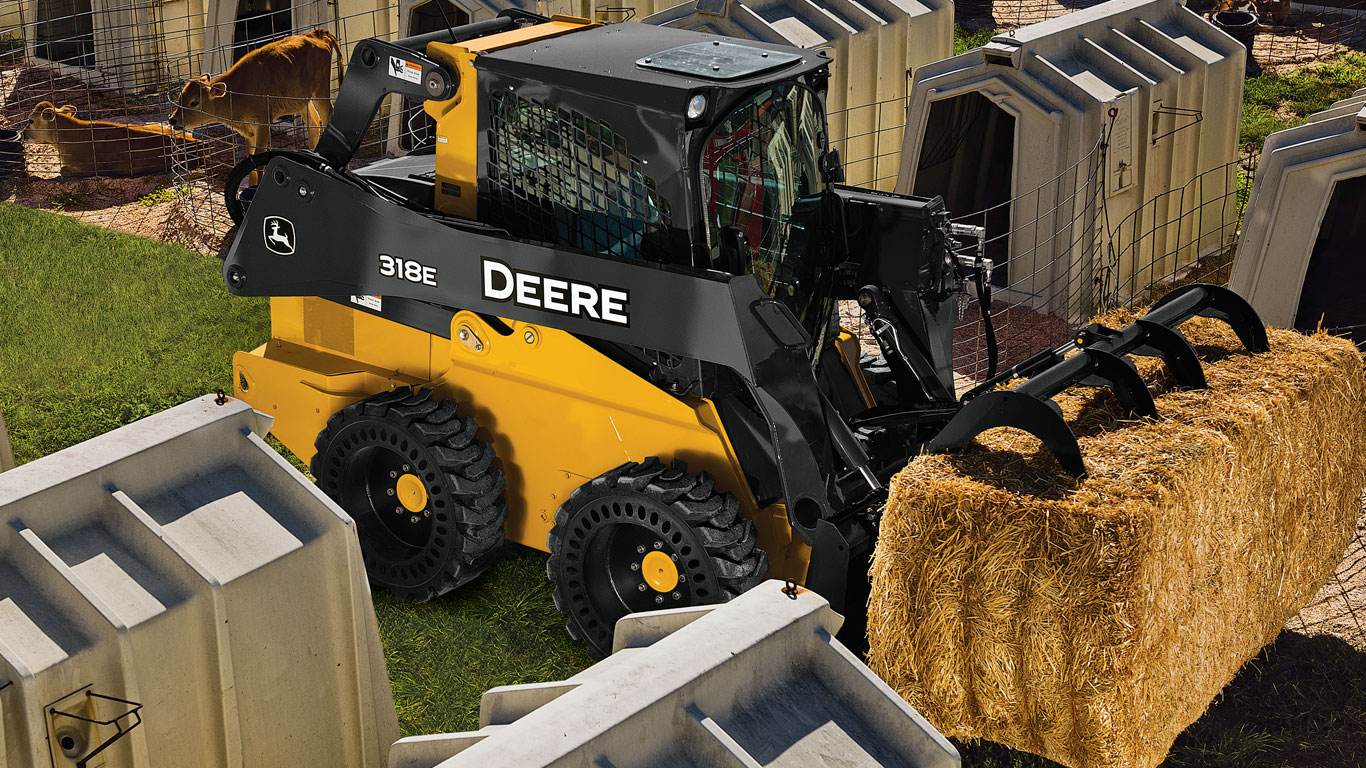 Une chargeuse à direction différentielle John Deere équipée de l’accessoire pinces pour rebuts transporte de la paille.